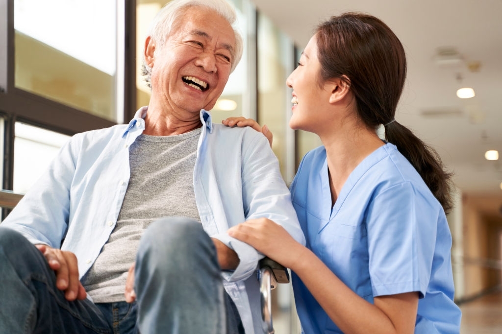 young friendly asian female caregiver talking chatting to happy senior man in hallway of nursing home